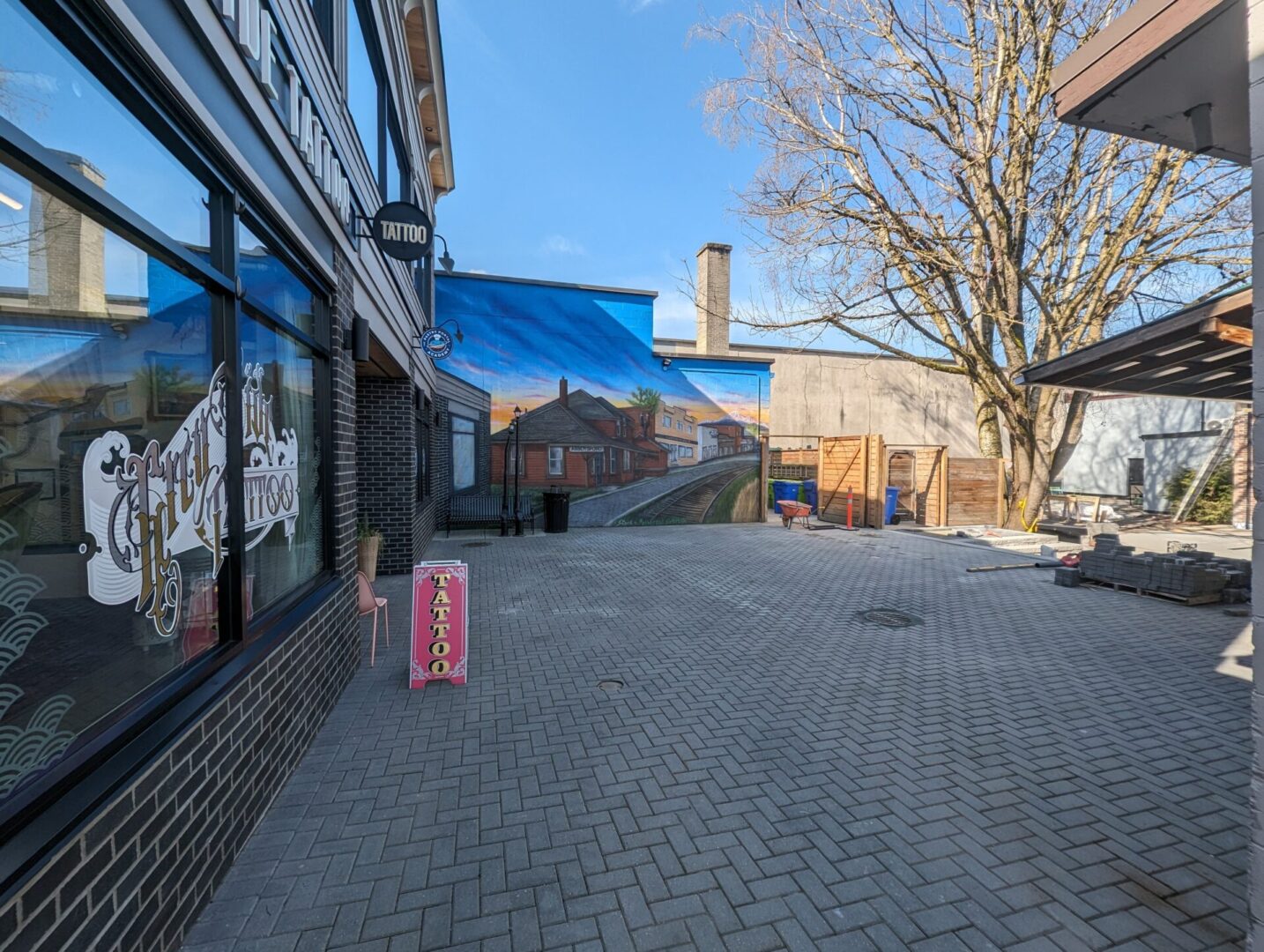 A vibrant street mural on a building wall next to a tattoo shop, featuring a serene landscape scene, viewed from a paved courtyard under bright daylight.